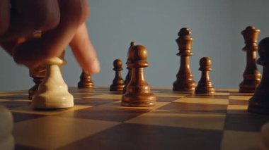 El, satranç tahtasının üzerinde stratejik olarak hareket eden beyaz piyon. Etrafı koyu ahşap satranç taşlarıyla çevrili. Sahne, oyun oynamanın yoğunluğunu ve odak noktasını yakalar. Karar verme becerisini sembolize eder.