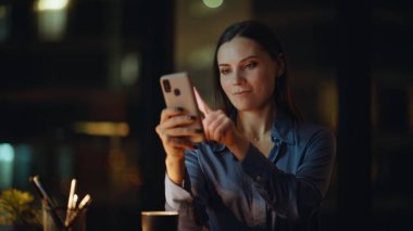 Gece iş yerinde cep telefonuyla internette sörf yapan neşeli bir kadın. Gülümseyen iş kadını ofisteki telefon uygulaması ile online alışveriş yapıyor. Akıllı telefon oturma masasında mesajları okuyan esmer bir kadın..