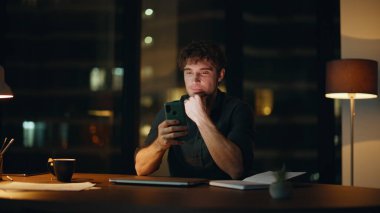 Kapalı dizüstü bilgisayarla karanlık bir iş yerinde cep telefonuna mesaj atan mutlu gece çalışanları. Gülümseyen adam, lamba ışığı altında akıllı telefondan mesaj atıyor. Rahat iş adamı geceleri cep telefonunda e-posta okuyor..