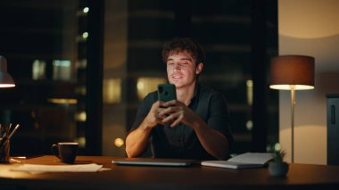 Çalışanların gece ofisinde lamba ışığı altında akıllı telefon mesajları. Gülümseyen işadamı, bankacılık telefonu uygulaması finansal geliri tatmin etti. Mutlu yönetici gece geç saatlerde iş yerinde cep telefonu mesajlarını okuyor.