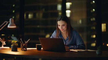 Kadın serbest çalışan, gece ofisindeki web kamerası laptopuyla konuşuyor. Gülümseyen iş kadını bilgisayar kapağını lamba ışığında kapatmayı bitirdi. Mutlu son yöneticiler başarılı online pazarlıklardan memnun.