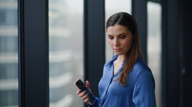 Pencere ofis binasında çalışan bir şirket çalışanı akıllı telefonu yakın çekimde tutuyor. Kendine güveni tam bir iş kadını. Şehir manzarasının keyfini çıkarıyor. Çalışma alanı telefonlu. İş planlama üzerine düşünen güzel bir kadın.