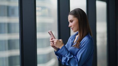 Ofisteki panoramik pencerede akıllı telefona mesaj atan zarif bir bayan. Gülümseyen esmer kadın iş yerindeki cep telefonu mesajları modern binanın içinde kırılıyor. Mutlu iş kadını telefondaki bankacılık uygulaması.