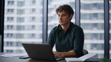 Motivasyonlu yönetici modern ofis alanındaki bilgisayarı daktilo ediyor. Panoramik pencerede kamerada gülümseyen profesyonel bir çalışan. Modern işyerinde şık bir iş adamı pozu.