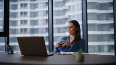 Ofiste oturan zengin bayan elinde kahve fincanı tutuyor. Gülümseyen iş kadını rahat koltuğunda rahatlıyor ve kariyer büyümesini düşünüyor. Kendine güvenen kadın yönetici dizüstü bilgisayarda başarılı olacağını tahmin ediyor..