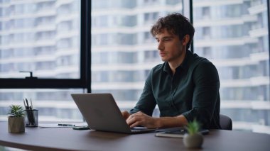 Modern ofis alanında dizüstü bilgisayarı daktilo eden kendine güvenen girişimci. Mutlu iş adamı kamerayı işaret ederek gülümsüyor ve işyerinde oturuyor. Bilgisayarda çalışan yakışıklı adam yöneticisi proje yaratıyor.