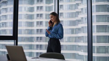 Gülümseyen yönetici cep telefonuyla modern şirket ofisini konuşuyor. Şehir manzaralı panoramik pencereden akıllı telefonu arayan mutlu iş kadını. Güzel kadın çalışanlar arkadaşça telefon konuşmaları yapar..