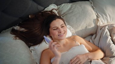 Uyanmış bir kadın cep telefonuyla konuşuyor. Beyaz yatak örtüsünün üstünde dinleniyor. Üst manzara. Gülümseyen esmer, sabah uyandıktan sonra akıllı telefondan arıyor. Evde cep telefonuyla konuşan uzun saçlı neşeli bayan..