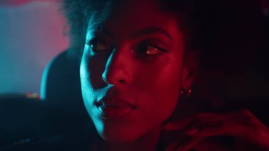 Portre kıvırcık saçlı model. Koyu renk araba pozu veriyor. Şehir ışıklarını yakıyor. Kırmızı neonun altında parlayan güzel Afro-Amerikan kadın. Ciddi kadın otomobil yolcusu tek başına pencereye bakıyor..
