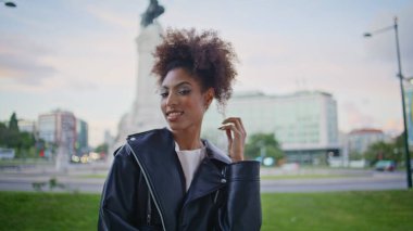 Kıvırcık kadın kamerada gülümsüyor bulutlu şehir caddesinde dikiliyor. Buklelerle oynayan mutlu Afrikalı Amerikalı kız portresi. Kaygısız moda kadını şehirde yalnız poz veriyor. Deri ceket giyiyor..