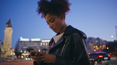 Deri ceketli, gece sokakta mesajlaşan Afrikalı Amerikalı bir kadın. Çekici kıvırcık saçlı bayan sokak lambalarının önünde cep telefonuna mesaj atıyor. Kendine güvenen esmer cep telefonu uygulaması.