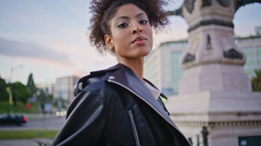 Deri ceketli şık model şehir meydanında duruyor. Metal parmaklıklara dayanan Afro-Amerikan kadın şehir mimarisi. Şık kıvırcık saçlı bayan tarihi caddede rahatlayan kameraya bakıyor.