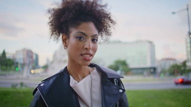 Afro-Amerikan model kamera canlı kentsel alanda yakın çekim yapıyor. Şehirli deri ceketli kıvırcık saçlı kadın. Moda makyajlı, güzel bir bayan kasabanın merkezinde poz veriyor..
