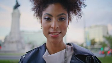 Şık bir portre modeli, kentin arka planında kendine güvenen bir kamera gibi görünüyor. Deri ceketli gülümseyen Afro-Amerikan kadın modern caddede ayakta duruyor. Şehre yakın poz veren kıvırcık saçlı mutlu moda kız..