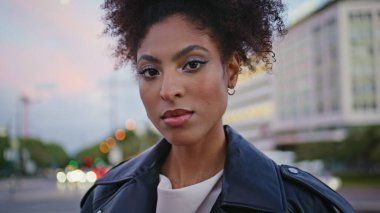 Kentin alacakaranlığında deri ceketle poz veren kendine güvenen kadın portresi. Muhteşem Afro-Amerikan kadın, kameralar kasabanın ışıklarını kapatıyor. Moda kıvırcık saçlı manken kız şehir merkezinde tek başına duruyor..