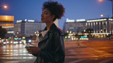 Esmer kadın gece aydınlanmış caddede cep telefonuna yakın çekim mesajı atıyor. Deri ceket giymiş kendine güvenen Afro-Amerikan kadın. Karanlık şehir manzarasında akıllı telefon gibi görünüyor. Kıvırcık kadın gece hayatının tadını çıkar.