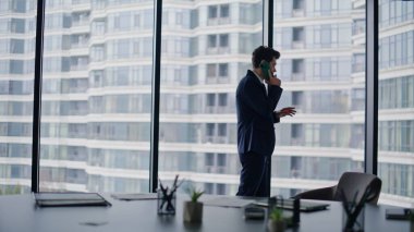 Şirket ofisindeki cep telefonuyla iş görüşmeleri. Zarif iş adamı panoramik pencerenin yanında akıllı telefondan konuşuyor. İşyerinde başarılı bir yönetici cep telefonuyla istemciye danışır.