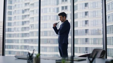 Kendine güvenen lider panoramik ofis penceresinde duruyor ve iş başarısını düşünüyor. Şık bir işadamı yüksek binalara bakıyor. Yakışıklı yönetici akıllı telefon düşünüp taşınma projesi tutuyor..