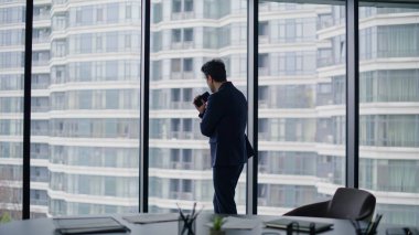 Düşünceli iş adamı ofis penceresinin yanında durup şehir manzarası izlemeyi düşünüyor. Akıllı telefon tutarak profesyonel düşünme stratejisi. Kararlı yönetici önemli kararlar alıyor.