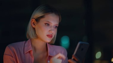 Gülümseyen iş kadını, gece ofisinde cep telefonunu okuyor. Mutlu kadın, karanlık evde akıllı telefondan dostça bir mesaj alıyor. Pozitif sarışın cep telefonundan komik içeriği izliyor.