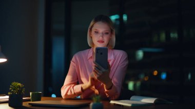 Gece ofisinde gülümseyen bir akıllı telefon. Yakın plan. Zarif bir iş kadını cep telefonunda bankacılık uygulamasına göz atıyor karanlık işyerinde oturuyor. Başarılı bir yönetici maaş telefonu bildirimi alıyor.
