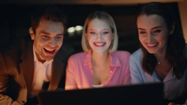 Gülümseyen arkadaşlar karanlık dairede dizüstü bilgisayara göz atıyorlar. Çevrimiçi içeriği izleyen mutlu gençler gece düşüncelerini paylaşıyorlar. Neşeli takım arkadaşları bilgisayar görünümlü takım projesinin keyfini çıkarın..