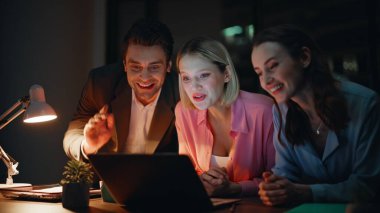 Gülümseyen çalışanlar gece iş yerinden arıyorlar. Neşeli meslektaşlar karanlık ofis alanında online toplantının keyfini çıkarıyorlar. Neşeli iş adamları el sallayarak laptopun web kamerasını selamlıyorlar.