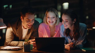 Amigo takım arkadaşları gece iş yerinde iletişim kuruyor ve dizüstü bilgisayarı yakından izliyorlar. Çevrimiçi proje başarısını tartışan gülümseyen meslektaşlar. Mutlu iş arkadaşları ofiste dostça sohbetin keyfini çıkarırlar..