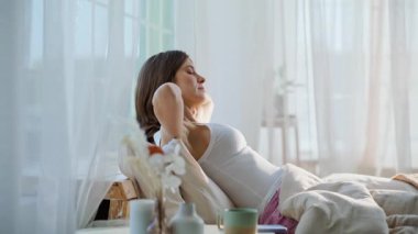 Yumuşak yastıklarda oturan kollarını esneten uykulu kız. Gülümseyen rahat kadın rahat beyaz yatak odasında tek başına cep telefonu kullanıyor. Mutlu sakin esmer evde romantik cep telefonu mesajları okuyor..