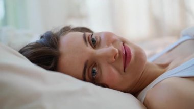 Kamerada gülümseyen, rahat ev yataklarında uzanan uykulu kız. Mutlu güzel bir kadın sabahın erken saatlerinde rahat bir dairede uyanıyor. Neşeli esmer, yatak odasında ferahlığın keyfini çıkarıyor..