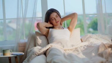 Ev yatağında kollarını uzatan uyanık bir kadın. Rahat genç bayan, yumuşak yataklarda oturup akıllı telefon kullanıyor. Gülümseyen esmer, sakin bir hafta sonunun keyfini çıkarıyor..