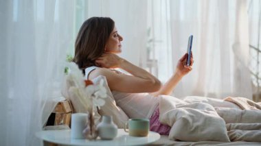 Uyanık kız rahat beyaz yatakta akıllı telefon gibi görünüyor. Gülümseyen çekici kadın sabah kahvesini yudumlarken cep telefonu haberlerini sunuyor. Kaygısız esmer, sakin bir hafta sonunun keyfini çıkarıyor..