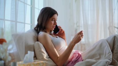 Cep telefonuyla konuşan sağlıksız bir kadın ev yatağında sıcaklığı ölçüyor. Sağlıksız esmer kadın yastıkların üzerinde yatan doktorla telefonda görüşüyor. Hasta kadın akıllı telefon ile hastalıkları tartışıyor..