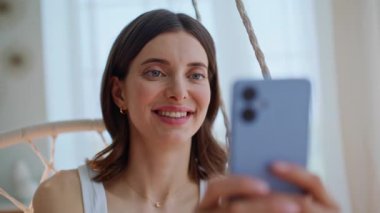 Akıllı telefonu elinde tutan neşeli bayan evde ip koltukta oturuyor. Yakın çekim seçici odak noktası. Gülümseyen esmer, ışık odasında cep telefonu mesajı okuyor. Sabah apartmanında cep telefonuna mesaj atan güzel bir kadın.