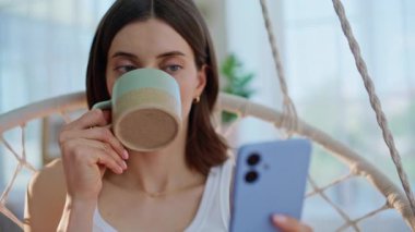 Ev hanımı akıllı telefondan mesaj okuyor rahat rahat asılı sandalyeye yakın oturuyor. Rahatlamış genç bir kadın sabah kahvesini içerken cep telefonu gibi görünüyor. Hafif apartmanda rahat esmer, cep telefonlu..