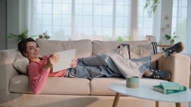 Oturma odasındaki kanepede uzanmış tablet izleyen bir kadın. Gülücükler saçan genç bir bayan, rahat bir ev kanepesinde, bilgisayarında medya içeriğini inceliyor. Rahat esmer kız internetten alet kullanarak alışveriş yapıyor..