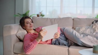 Rahat, esmer, göz gezdirme tableti rahat kanepe oturma odasında yakın çekim yatıyor. Gülücükler saçan genç bir kadın, içeriği izlerken sekme bilgisayarında kanepede dinleniyor. Dijital cihaza bakan mutlu ev hanımı.