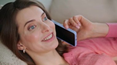 Fotoğrafçı kız telefonda kanepe yastıklarına bakarak gülüyor. Mutlu kadın, haber paylaşma planlarını cep telefonu konuşmalarında ev kanepesinde tartışıyor. Gülümseyen esmer, akıllı telefon iletişiminin keyfini çıkar..