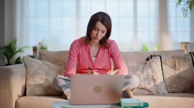 Sanal sınıfta okuyan bir kadın evdeki kanepede bilgi topluyor. Zeki esmer kız dizüstü bilgisayarda eğitim videosu izliyor. Not defteri yazıyor. Özenli öğrenci. Kanepede online öğretmen dinle..