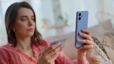Kadının kredi kartı bilgilerini akıllı telefon uygulamasına girmesi ev koltuğu kapanışında online alışveriş siparişi vermesi. Gülümseyen ev kadını internetten cep telefonu uygulaması kullanarak alışveriş yapıyor. Kanepede telefon ödemesi yapan mutlu kadın.