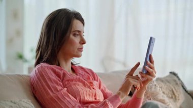 Aşırı neşeli bir kadın akıllı telefon ekranında oturuyor, kanepeye yakın oturuyor. Heyecanlı esmer, evde cep telefonuyla rahatlarken harika haberler okuyor. Mutlu bayan şaşırdı. Cep telefonunu izleyerek kazandı..