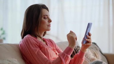 Dairedeki kanepe kapanışında cep telefonuyla kayıt yaptıran bir kız. Gülümseyen esmer, rahat koltukta oturmuş akıllı telefon sosyal medya izliyor. Cep telefonu mesajlarına bakan güzel kadın.