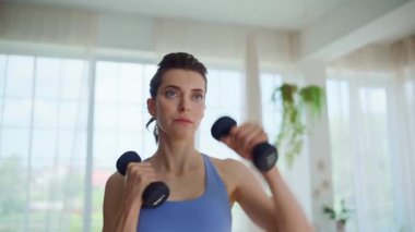 Oturma odasında dambıllarla egzersiz yapan bir kadın. Evde ağırlık kaldırma eğitimi alan motive olmuş genç bir bayan. Odaklanmış sporcu kadın vücut gücü için sabah egzersizi. Ağırlık kaldırma kavramı