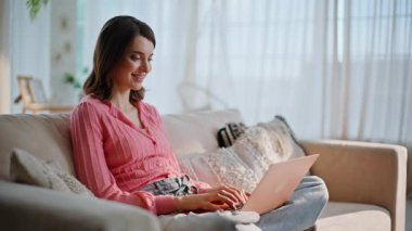 Rahat bir bayan rahat bir dairede klavye dizüstü bilgisayar yazarak online proje üzerinde çalışıyor. Rahat koltukta bacak bacak bacak üstüne atmış oturan genç esmer e-postaları cevaplıyor. Gülümseyen kadın web iletişiminden hoşlanıyor.