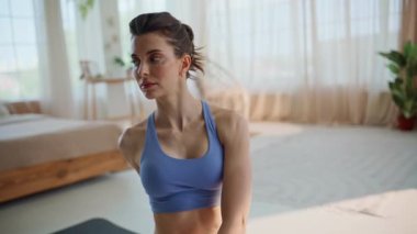 Güzel bir atlet, spor minderinin önünde otururken kaslarımı esnetiyor. Parlak dairede sağlıklı bir vücudu ısıtan sportif bir kadın. Motive olmuş esmer kadın modern oturma odasında pilates egzersizi yapıyor..