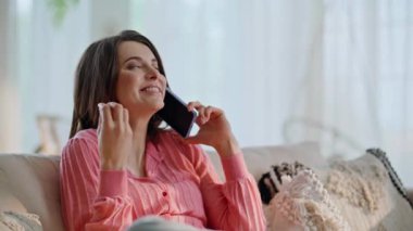 Neşeli esmer, konuşan cep telefonlu rahat kanepe kapanışı. Gülümseyen kadın dairede akıllı telefondan duygusal olarak harika haberleri tartışıyor. Güzel bayan cep telefonunu arıyor..