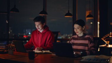 Modern çalışma ofisinde geç saatlere kadar çalışan ekip çalışanları. Gece işyerinde online projede işbirliği yapan iki gülümseyen yönetici. Dost canlısı meslektaşlar dizüstü bilgisayarlarla iş konuşuyorlar..