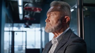 Modern şirket salonunda zarif bir takım elbiseyle duran düşünceli kıdemli işadamı. Gri saçlı şirket sahibi iş büyümesini düşünerek sakala dokunuyor. Ciddi bir yönetici hedefleri düşünüyor..