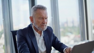 Ofis işyerindeki bilgisayarda çalışan finans müdürü. Yakın plan. Çalışma alanında dizüstü bilgisayar yazıp evrak okumakla meşgul kıdemli bir işçi. Kağıtları inceleyen sakallı iş adamı içeride yatırım sonuçları konusunda endişeli.