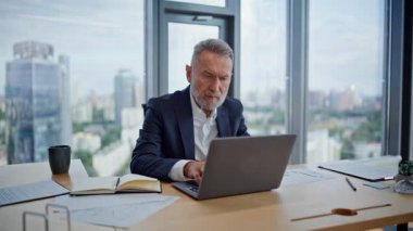 Modern iş yerinde dizüstü bilgisayar planlama projesine bakan olgun bir yönetici. Sakallı iş adamı, ışık ofisindeki bilgisayarda çalışıyor. Masada kağıt okuyan yaşlı girişimci gözatma uygulaması.